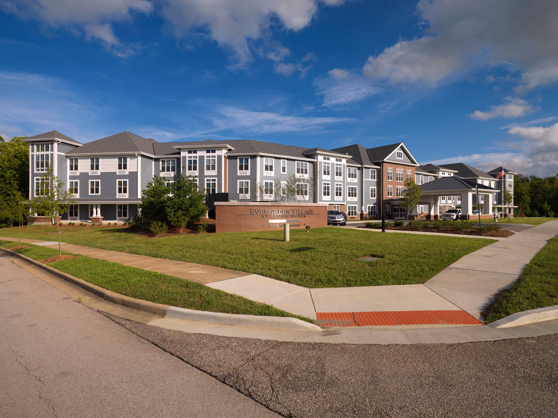 Exterior view of Evergreen Village at Bloomington, a large multi-story senior living facility with gray and white siding and brick accents, surrounded by green lawns and sidewalks under a partly cloudy blue sky.