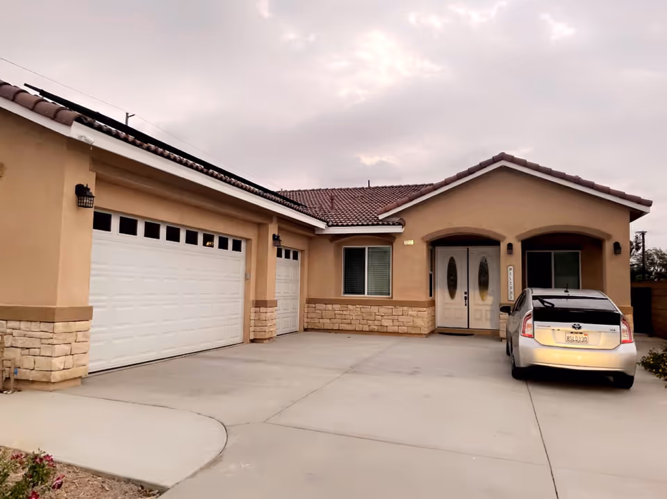 Exterior view of a single-story residential building with beige stucco walls and a tiled roof. The building features a double garage door, a smaller single garage door, a window with blinds, and a double front door with decorative glass panels. A silver car is parked in the driveway under an overcast sky.