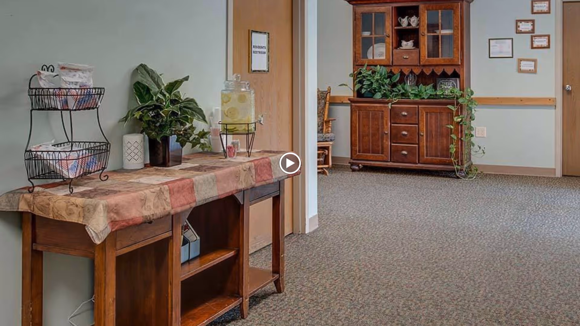 Carpeted interior common area with a wooden console table topped with plants and a beverage dispenser and a wooden hutch against the far wall.