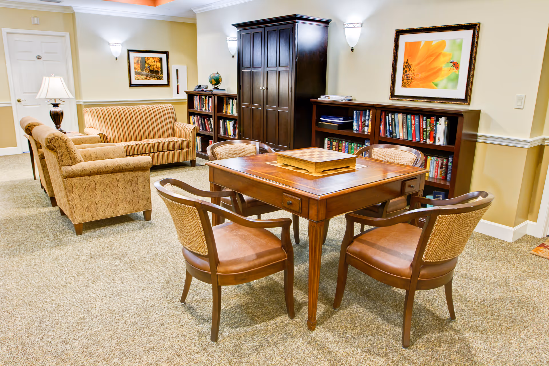 A cozy common area in a retirement facility featuring a wooden table with a chessboard on top, surrounded by four chairs. In the background, there are two bookshelves filled with books, a dark wooden cabinet, a striped sofa, and two patterned armchairs. The walls are decorated with framed pictures and wall-mounted lights.