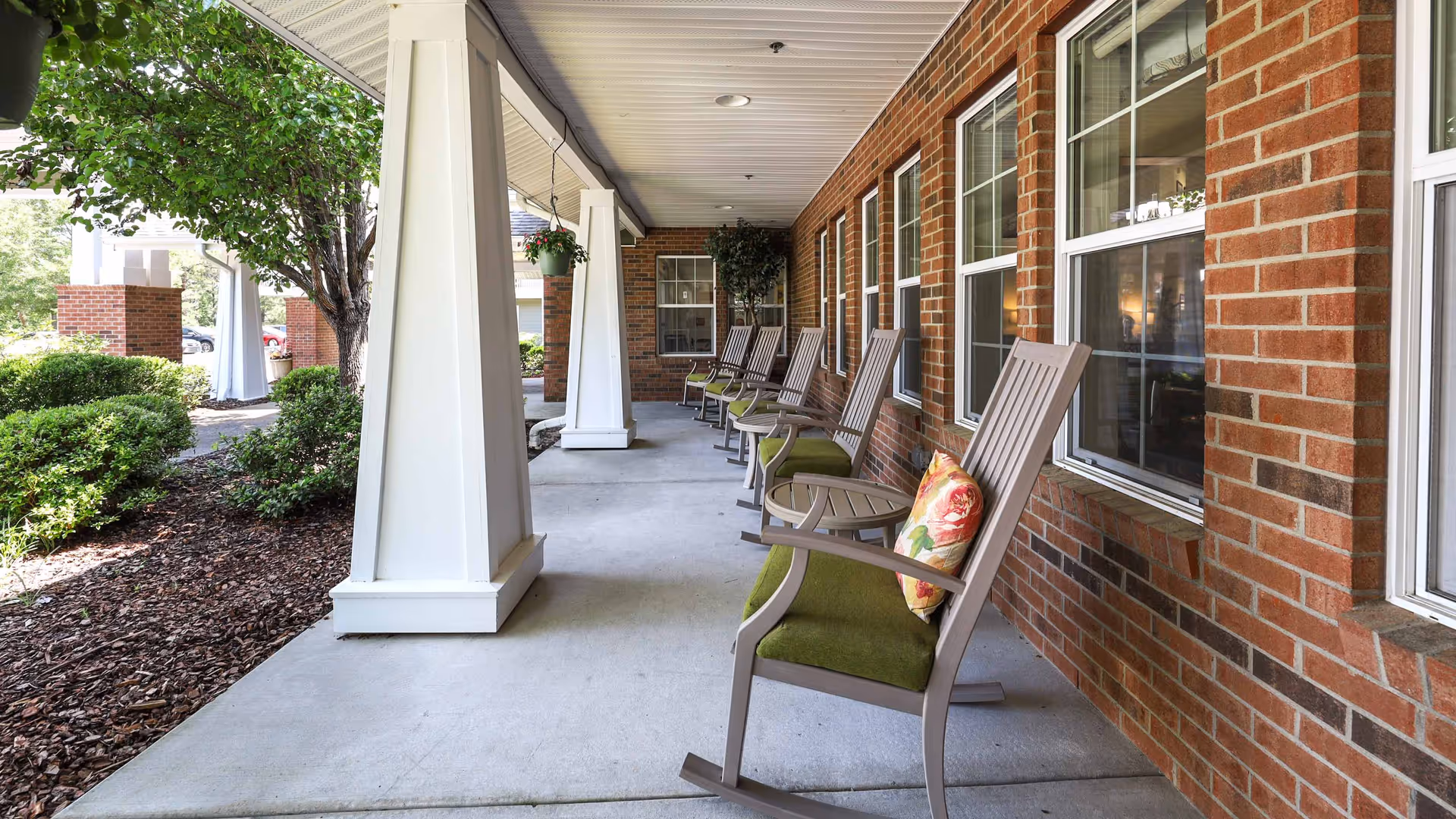 Covered outdoor porch area with a row of rocking chairs with green cushions and floral pillows along a brick wall with windows. The porch overlooks landscaped bushes and trees.