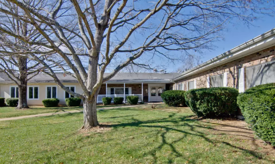 Exterior view of a single-story brick and siding building with multiple windows and a covered entrance. There is a large leafless tree in the grassy front yard along with trimmed bushes under a clear blue sky.
