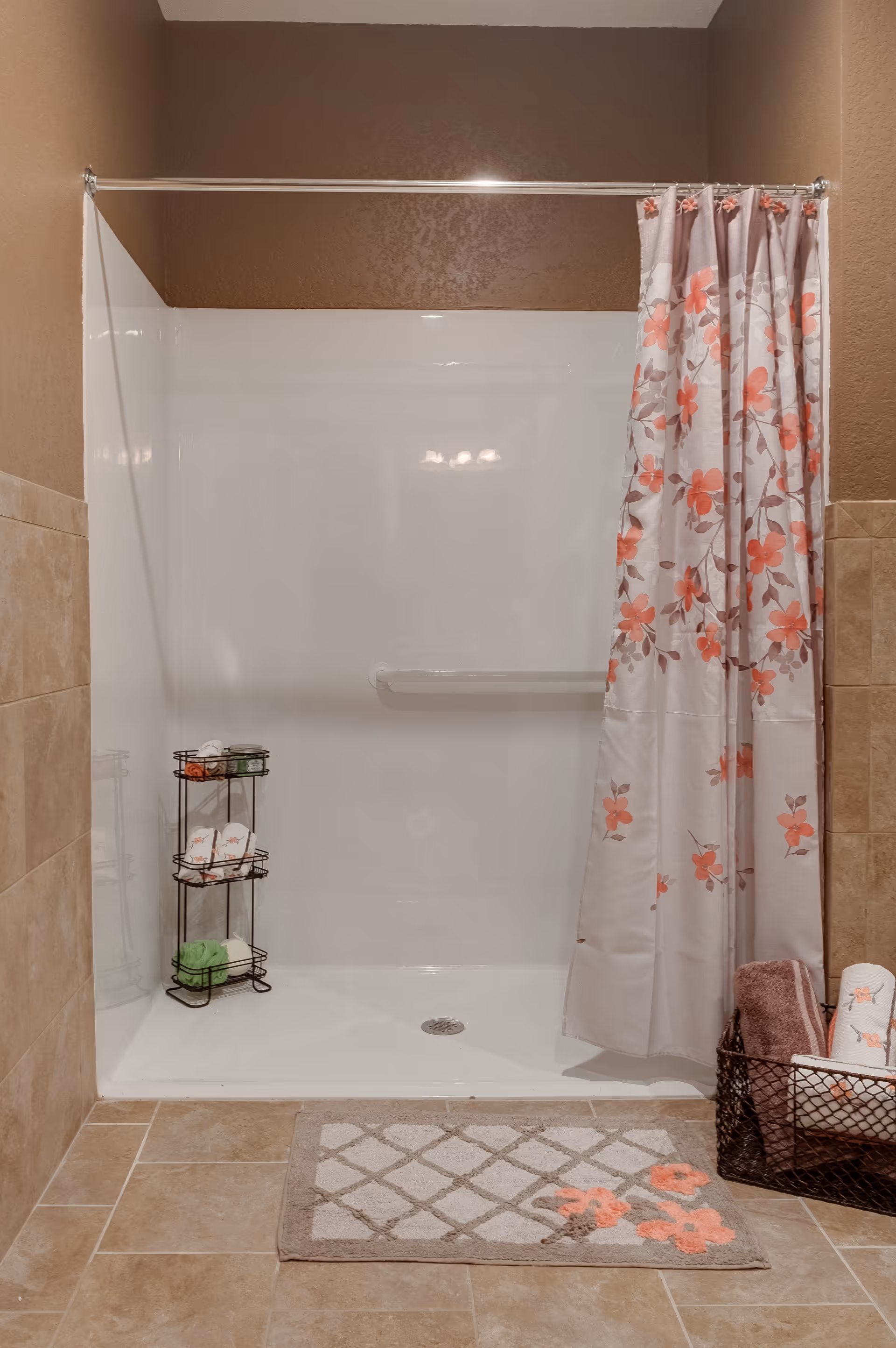 A clean shower area with beige tiled walls and floor. The shower has a white base and a floral shower curtain with pink and orange flowers. Inside the shower, there is a black metal rack holding toiletries and towels. Outside the shower, there is a beige and white patterned bath mat with orange flowers, and a black wire basket containing rolled towels.