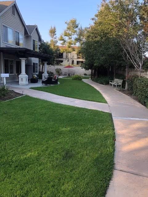 Curved concrete pathway winds through a green courtyard with benches, patio seating, trees, and a two-story residential building.