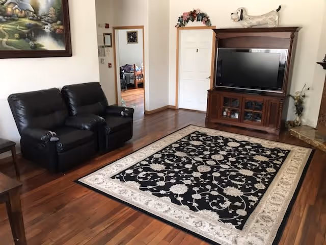 A living room with two black leather recliners on the left, a large patterned area rug in black and white on wooden flooring, a wooden TV cabinet with a flat-screen television, and a decorative dog figurine on top. There is a doorway leading to another room with a bed visible, and a painting on the wall above the recliners.