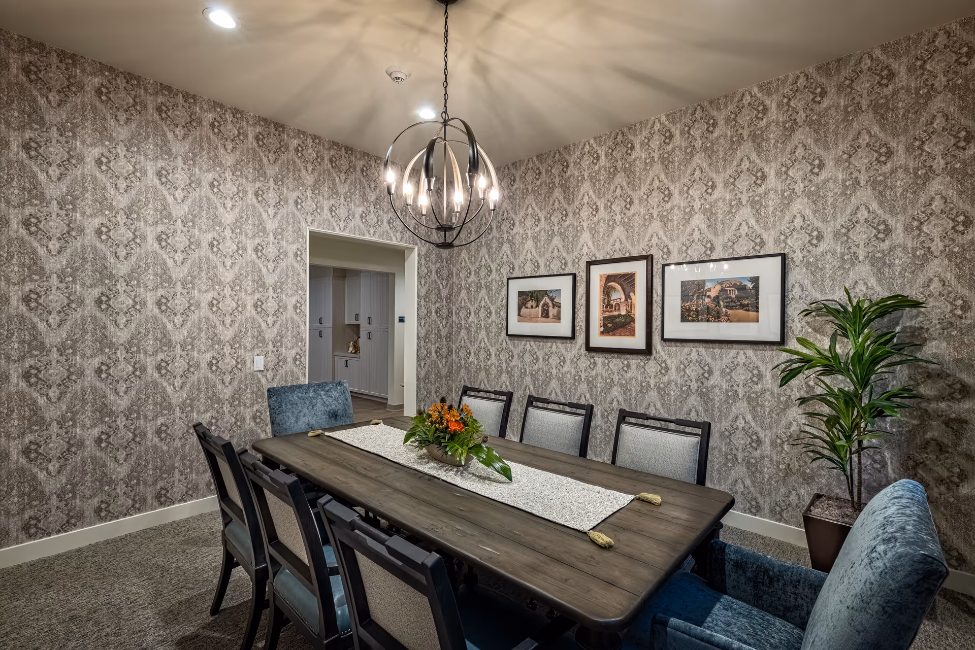 A dining room with a long wooden table surrounded by eight chairs, two of which are upholstered in blue fabric. The walls are covered with patterned wallpaper and decorated with three framed pictures. A modern chandelier hangs above the table, and a potted plant is placed in the corner near the wall.