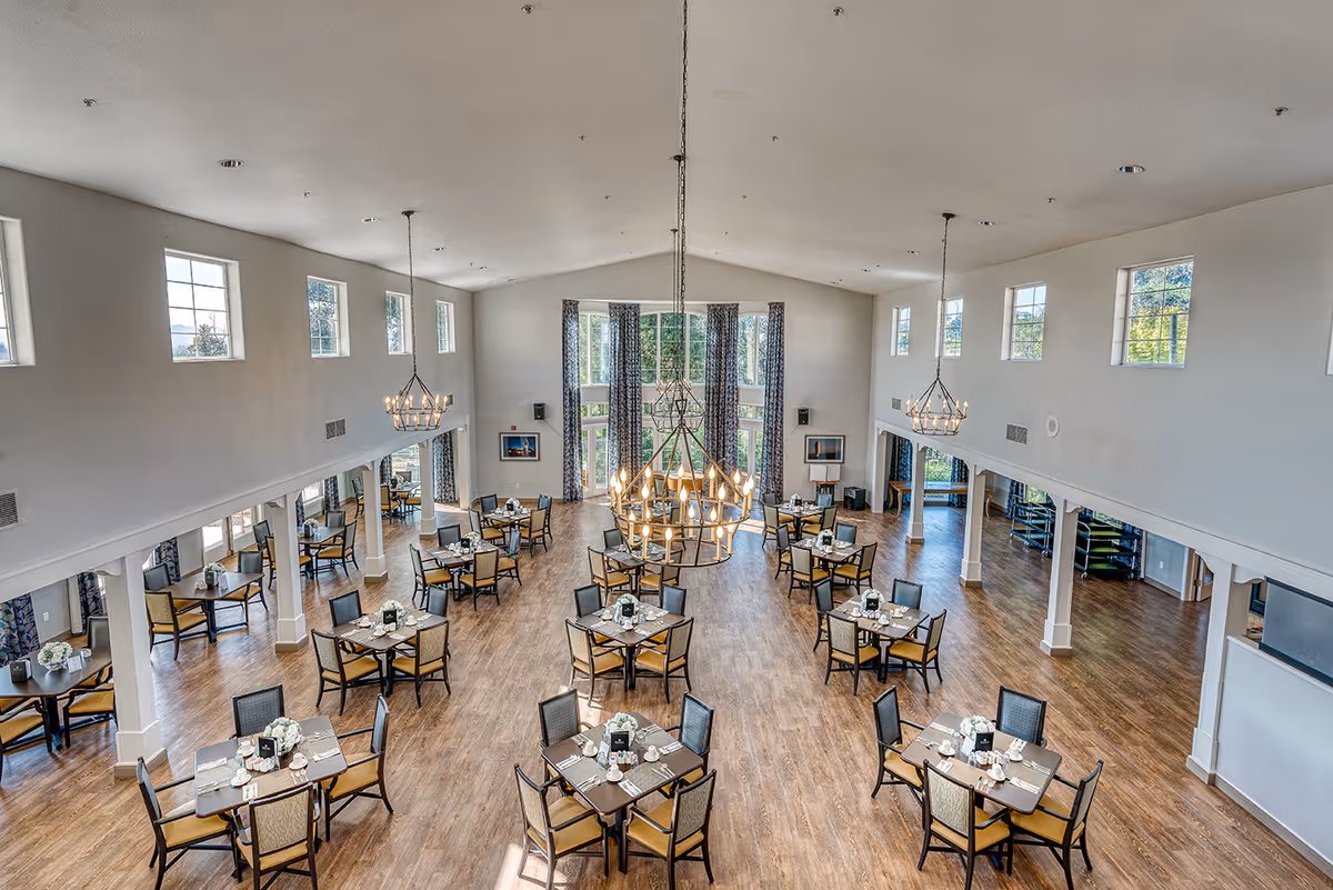 Spacious high-ceilinged dining room with multiple tables set for meals, large chandeliers, and tall windows.