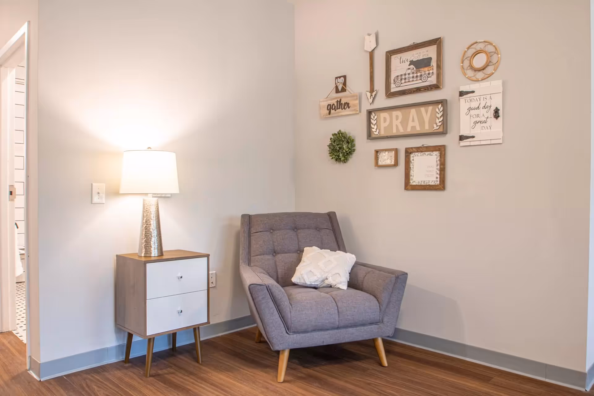 Cozy corner featuring a gray upholstered armchair, a mid-century side table with a lamp, and framed wall decor on a neutral wall.