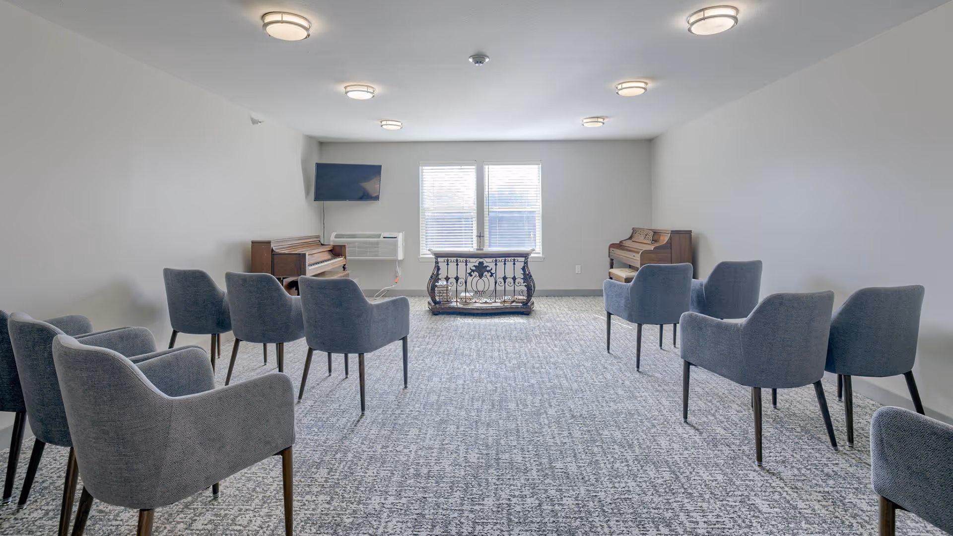 Bright common room with rows of gray upholstered chairs facing two pianos, a TV on the wall, and a window at the far end.