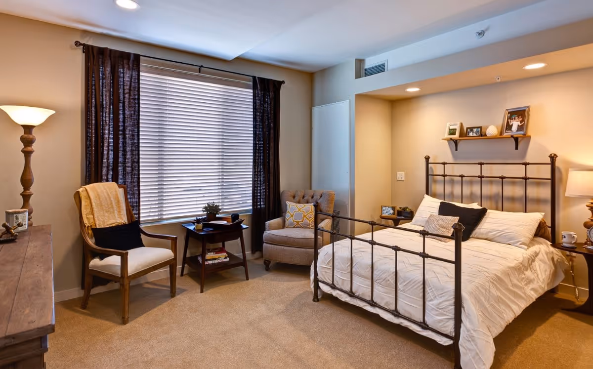 Well-lit furnished bedroom with a metal-framed bed, armchairs, side tables, and a large window with blinds.
