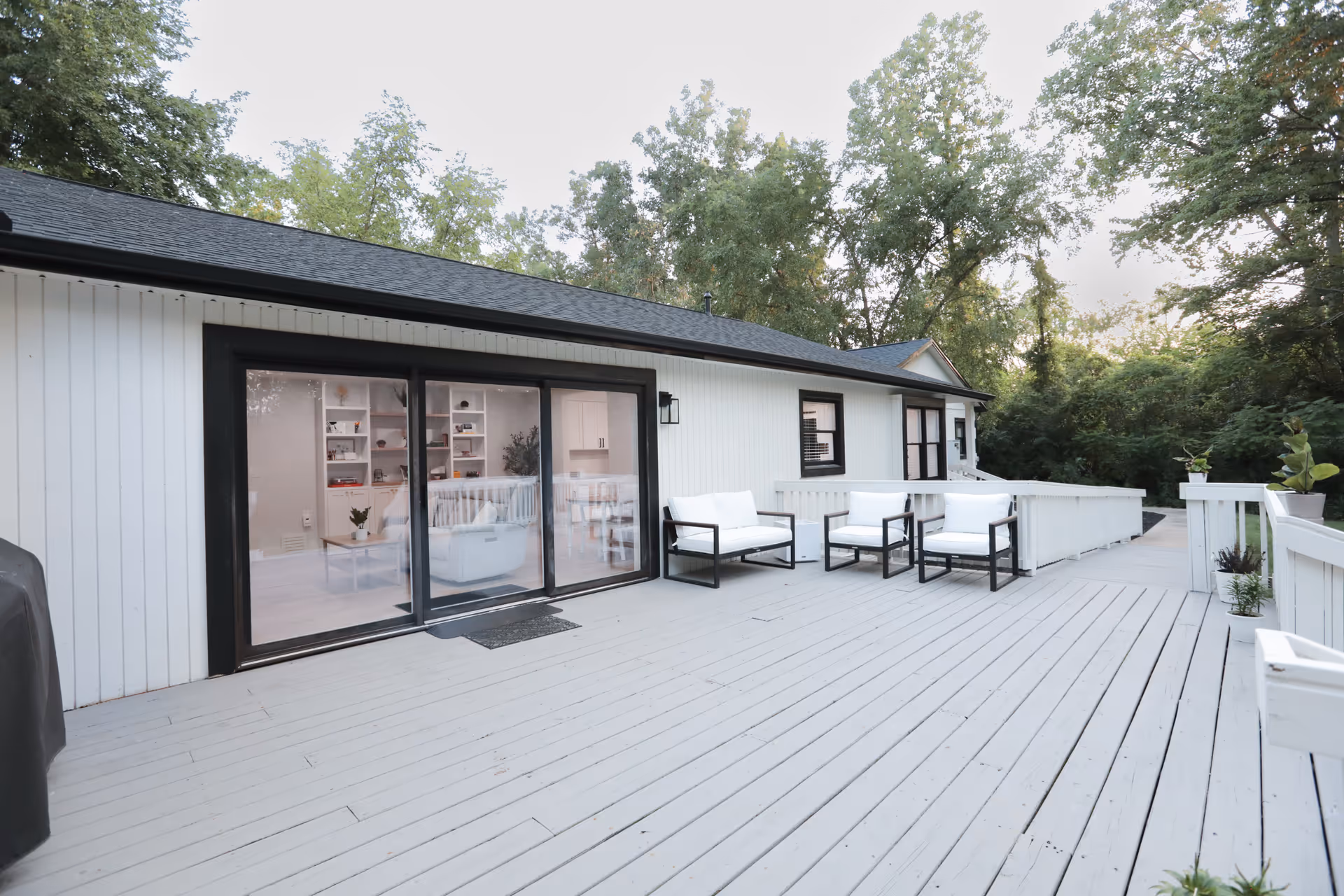 A spacious outdoor wooden deck attached to a white building with black trim. The deck features white cushioned chairs with black frames arranged near the sliding glass doors that lead inside. The area is surrounded by trees and greenery, creating a peaceful and natural setting.