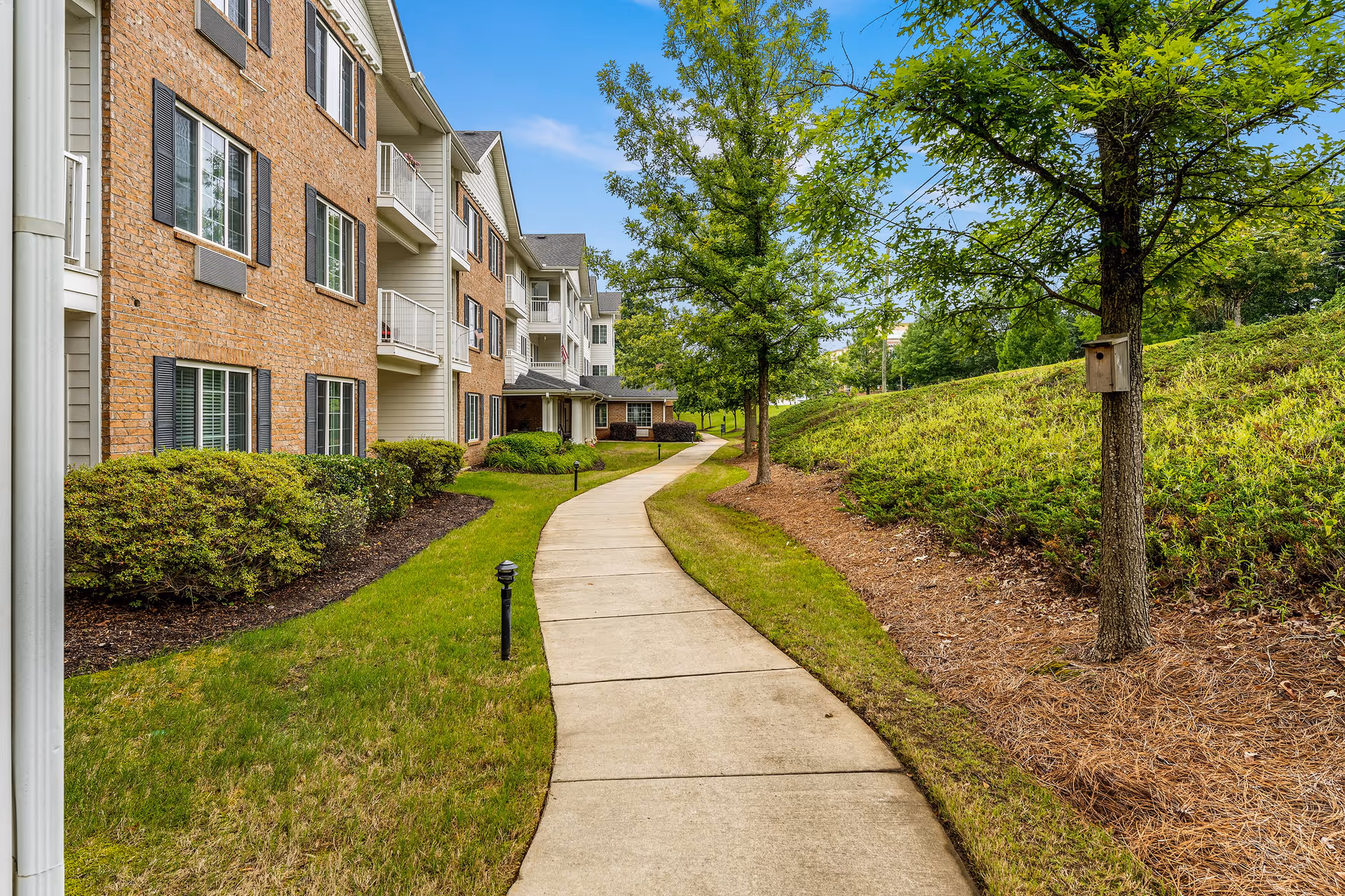 A winding concrete walkway runs alongside a multi-story brick and siding building with balconies. The path is bordered by neatly trimmed bushes and green grass on one side, and a grassy hill with trees and a birdhouse on the other side under a clear blue sky.