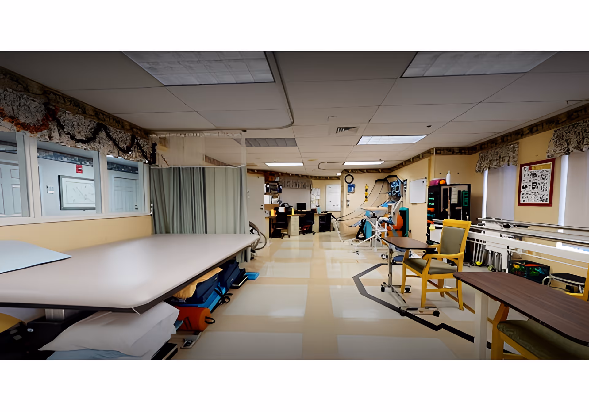 Interior view of a healthcare facility room with a large table on the left, chairs and tables on the right, medical equipment, and a work area with computers in the background. The room has a tiled floor, fluorescent ceiling lights, and windows with patterned valances.