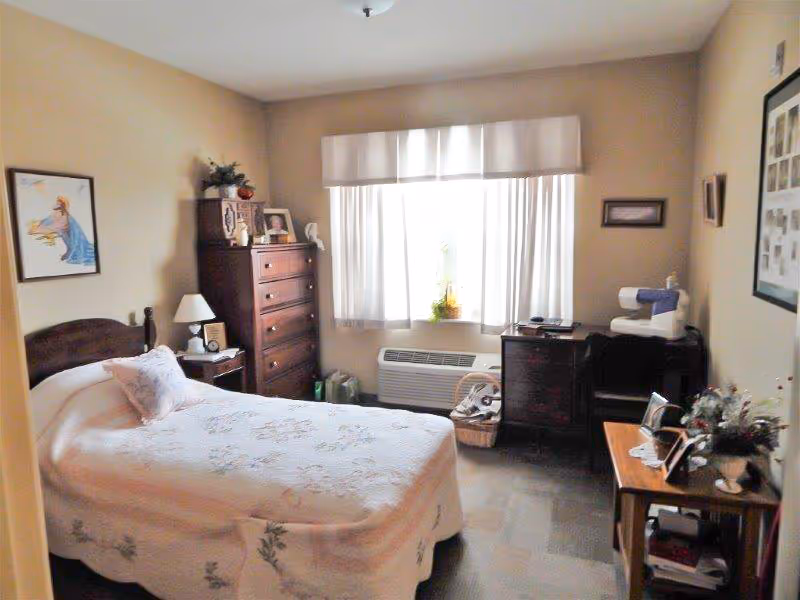 A cozy bedroom with a single bed covered in a white floral bedspread. There is a wooden nightstand with a lamp and clock beside the bed. A tall wooden dresser stands against the wall near a window with white curtains. A small desk with a sewing machine and other items is positioned under the window. The room has beige walls and carpeted flooring, decorated with framed pictures and plants.