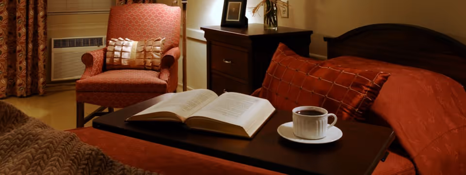 A cozy bedroom scene with a bed covered in a rust-colored bedspread and matching pillows. A dark wooden tray on the bed holds an open book and a white cup with a saucer filled with a hot beverage. In the background, there is a red patterned armchair with a decorative pillow, a wooden nightstand with a framed photo and a lamp, and a window with patterned curtains and an air conditioning unit.
