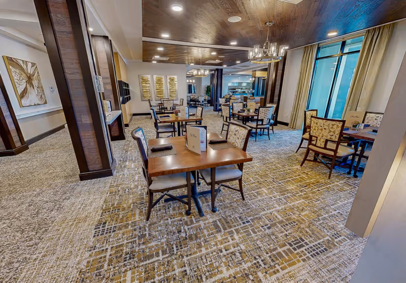 A spacious dining room with multiple wooden tables and cushioned chairs arranged neatly. The room features patterned carpet flooring, large windows with curtains, modern chandeliers, and decorative wall art. The overall ambiance is warm and inviting.