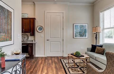 A cozy living room area with a light gray sofa adorned with dark and patterned cushions, a floor lamp beside it, and a small round coffee table with decorative items on a patterned rug. The room has wooden flooring, a white door in the center, a window with blinds on the right, and a small kitchen area with dark wooden cabinets visible on the left. The walls are decorated with framed artwork.