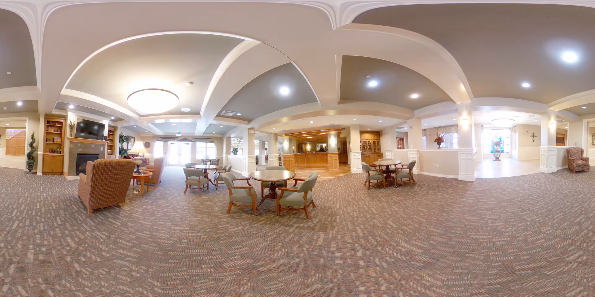 A spacious senior living community common area with carpeted floors, multiple round tables with chairs, comfortable armchairs, a fireplace with a mounted TV above it, and bright overhead lighting. The area has a warm and inviting atmosphere with beige walls, white ceiling beams, and decorative plants. There is a reception or service desk area in the background and large windows letting in natural light.