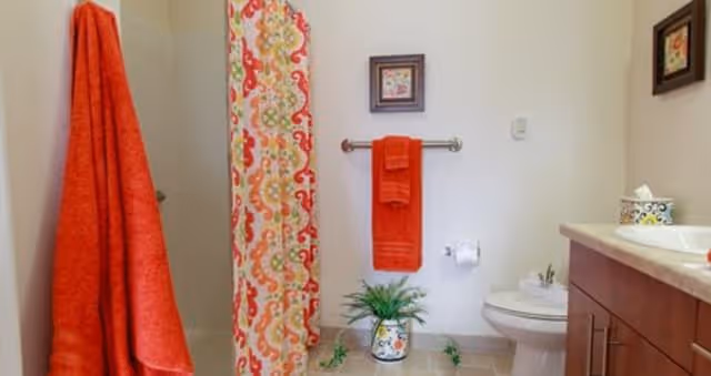 A bathroom with a white toilet, a wooden vanity with a sink, a colorful shower curtain with red, orange, and green patterns, and two orange towels hanging on silver towel racks. There is a small framed picture on the wall and a decorative plant in a patterned pot on the floor.