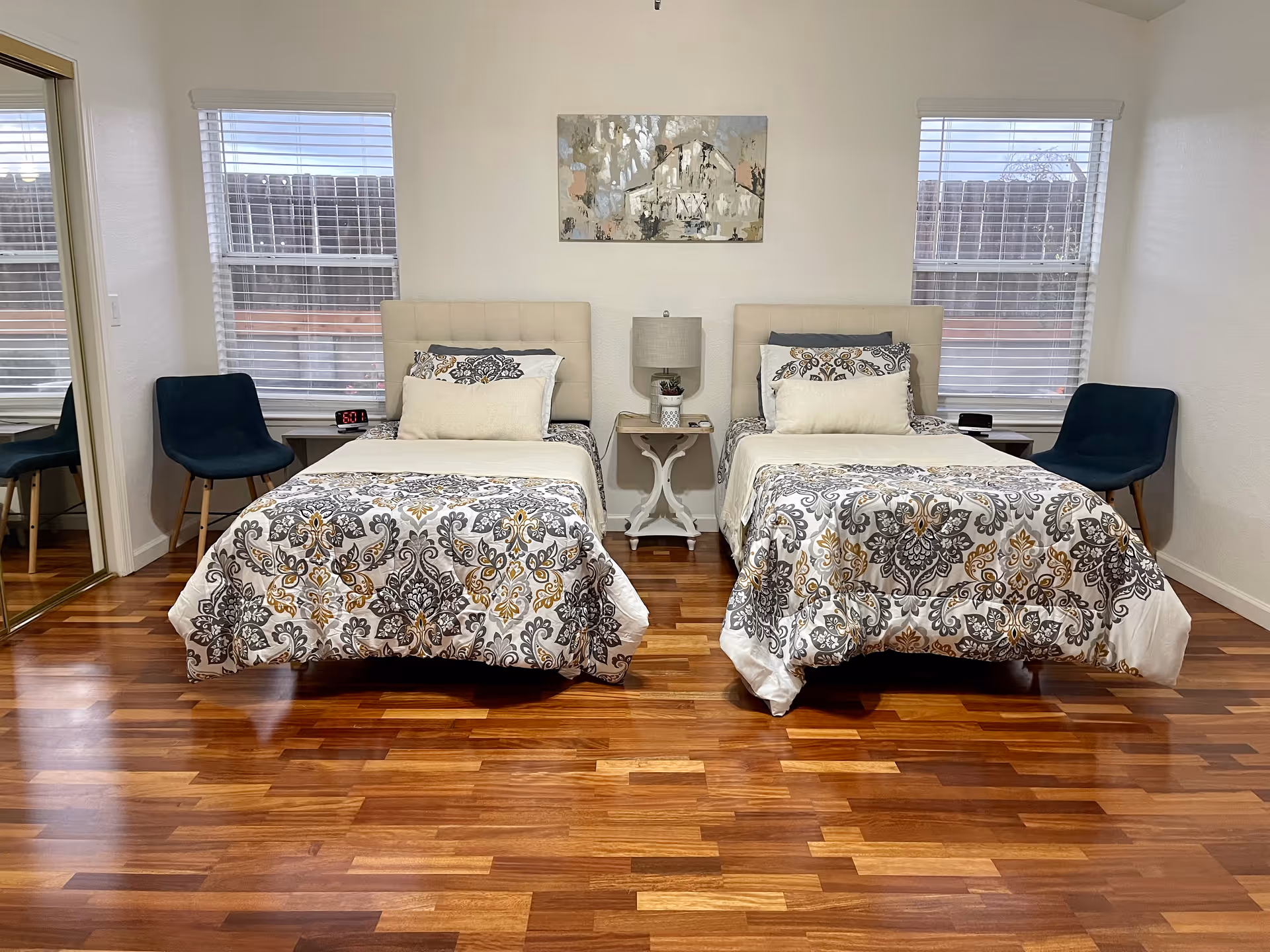 A bedroom with two single beds side by side, each with patterned bedspreads in shades of gray, white, and gold. Between the beds is a small white nightstand with a lamp and a small plant. There are two windows with blinds behind the beds, and two dark blue chairs placed near each window. The floor is wooden, and a large mirror is visible on the left wall.