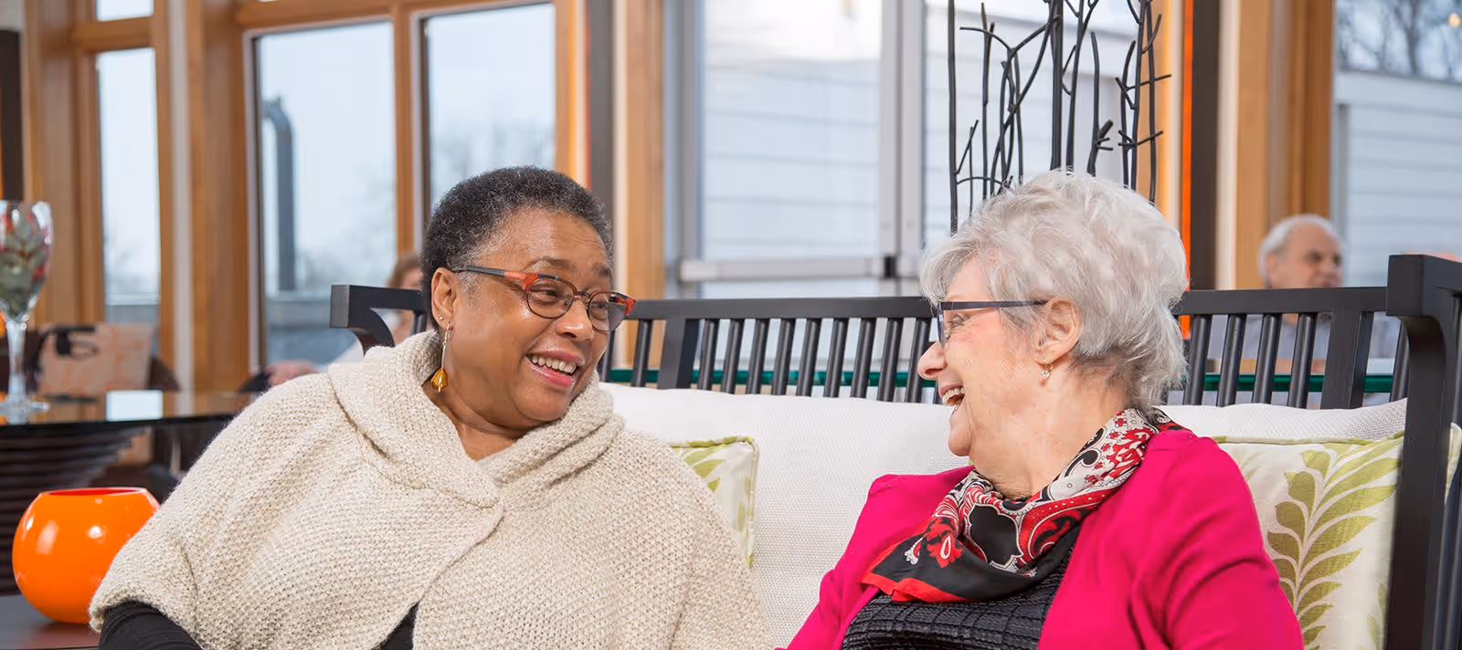 Two elderly women sitting on a cushioned bench indoors, smiling and engaging in a friendly conversation. The room has large windows with a view outside and decorative elements including a glass table with a vase and a metal coat rack behind them.