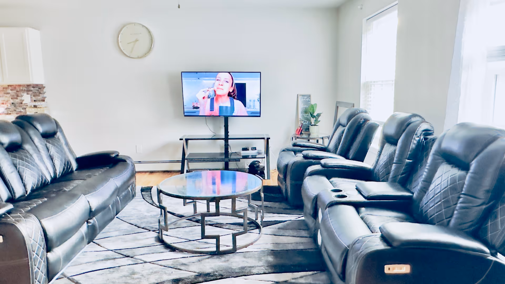 A bright living room with two black leather sofas facing each other, a round glass coffee table in the center, a flat-screen TV mounted on a stand showing a woman, a wall clock above the TV, and windows letting in natural light.