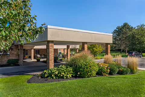 Exterior view of Elderwood at Hamburg facility showing a covered entrance with brick pillars, surrounded by well-maintained green lawn, shrubs, and trees under a clear blue sky.
