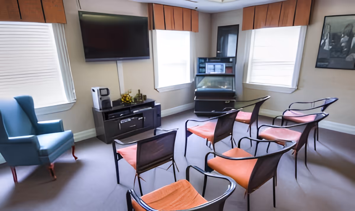 A small lounge area with six metal-framed chairs with orange cushions arranged in two rows facing a wall-mounted flat-screen TV. There is a blue armchair to the left near a window with blinds and valance, and a black cabinet with a coffee maker and decorative items below the TV. Two windows with white blinds and orange valances flank the TV, and a framed picture hangs on the right wall.