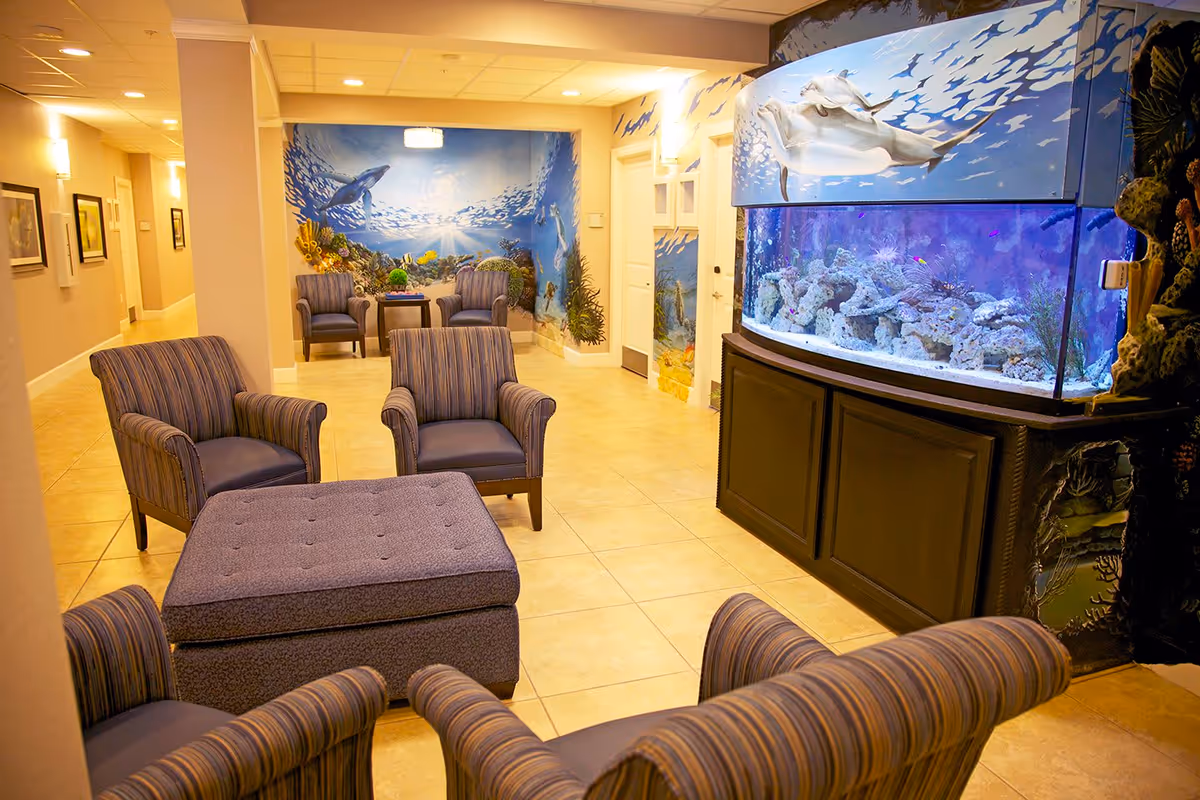 A cozy seating area in a senior living facility with four striped armchairs arranged around a large cushioned ottoman. In the background, there is a large aquarium with colorful fish and coral, and the walls are decorated with an underwater ocean mural featuring marine life such as a stingray and fish. The room has warm lighting and tiled floors.