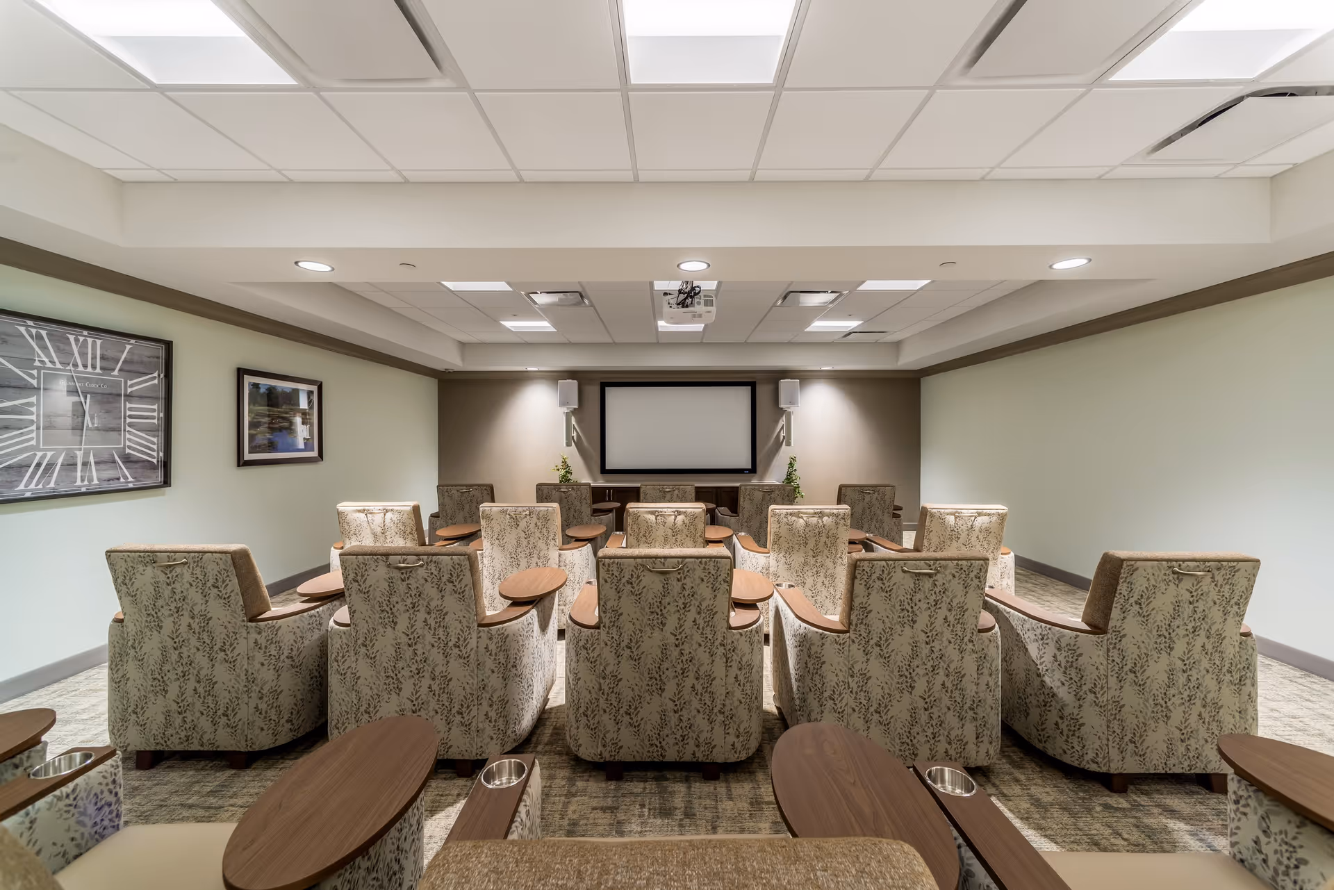 A small theater room with three rows of cushioned armchairs featuring floral upholstery and attached wooden side tables. At the front of the room is a large projection screen flanked by two wall-mounted speakers and two small plants on a wooden console. The ceiling has recessed lighting and a projector mounted in the center. The walls are painted light green and beige, with framed artwork on the left wall.