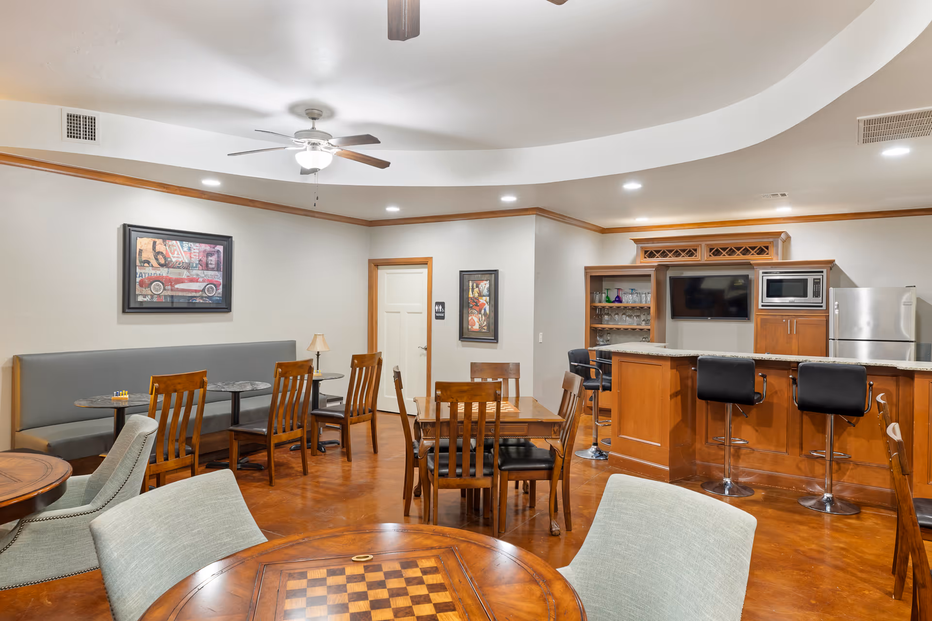 A well-lit common area in a senior living facility featuring multiple wooden tables and chairs, a built-in bar with granite countertop, bar stools, a microwave, and a refrigerator. The room has a ceiling fan, framed artwork on the walls, and a door with a restroom sign.