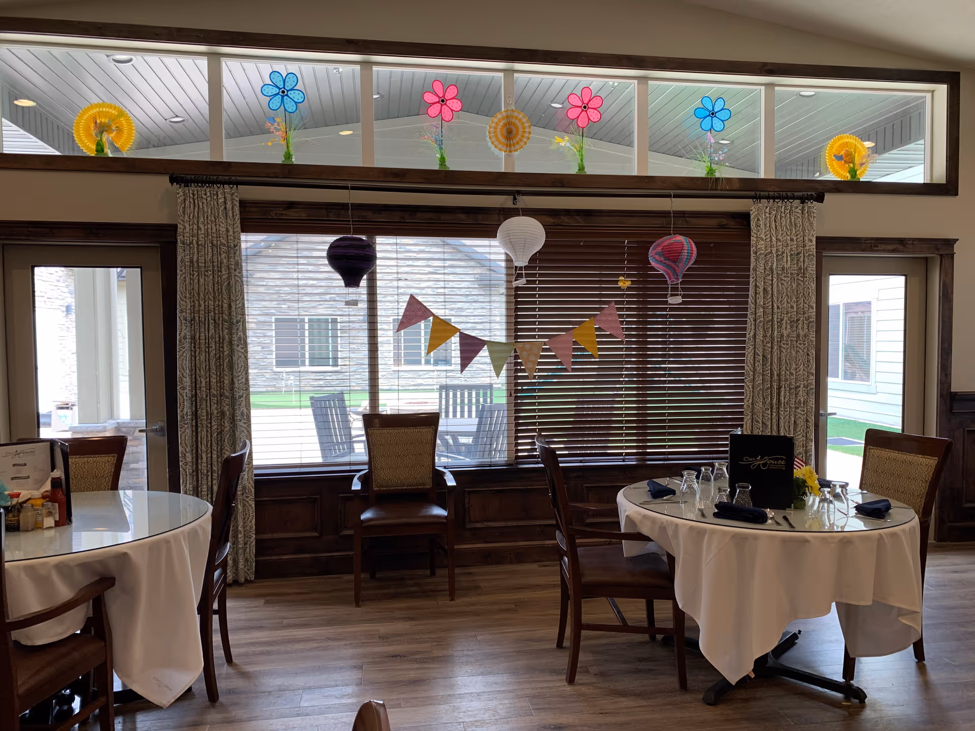 Dining area with round tables covered in white tablecloths, wooden chairs, and place settings including glasses and napkins. Large windows with blinds and colorful paper decorations such as flowers, hot air balloons, and a pennant banner hang in front of the windows. Outside, patio furniture is visible.