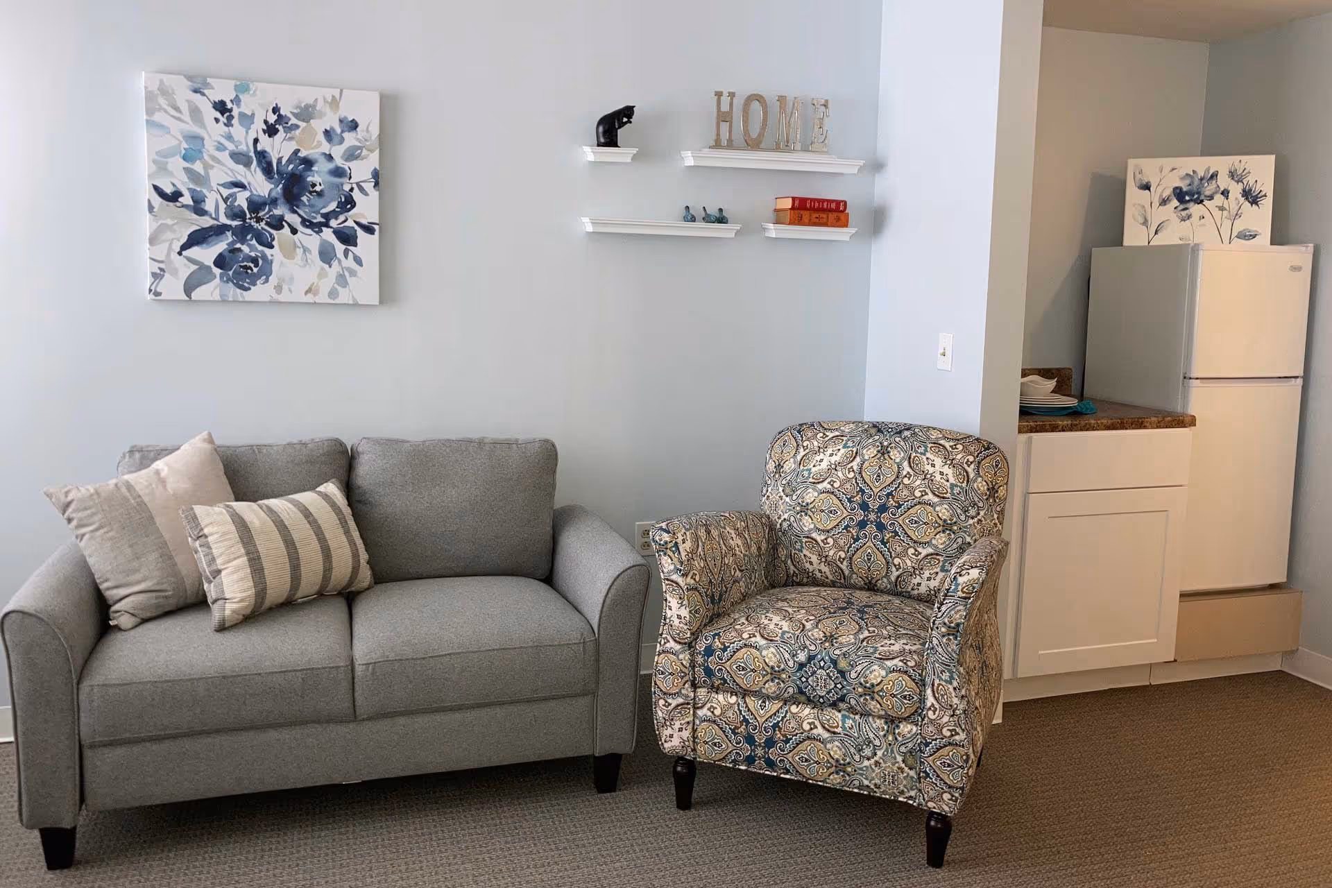 A cozy living area featuring a gray loveseat with two pillows, a patterned armchair, and a small kitchenette with a white refrigerator and countertop. The wall is decorated with a floral painting and three small white shelves holding decorative items and books, with the word 'HOME' displayed on the top shelf.