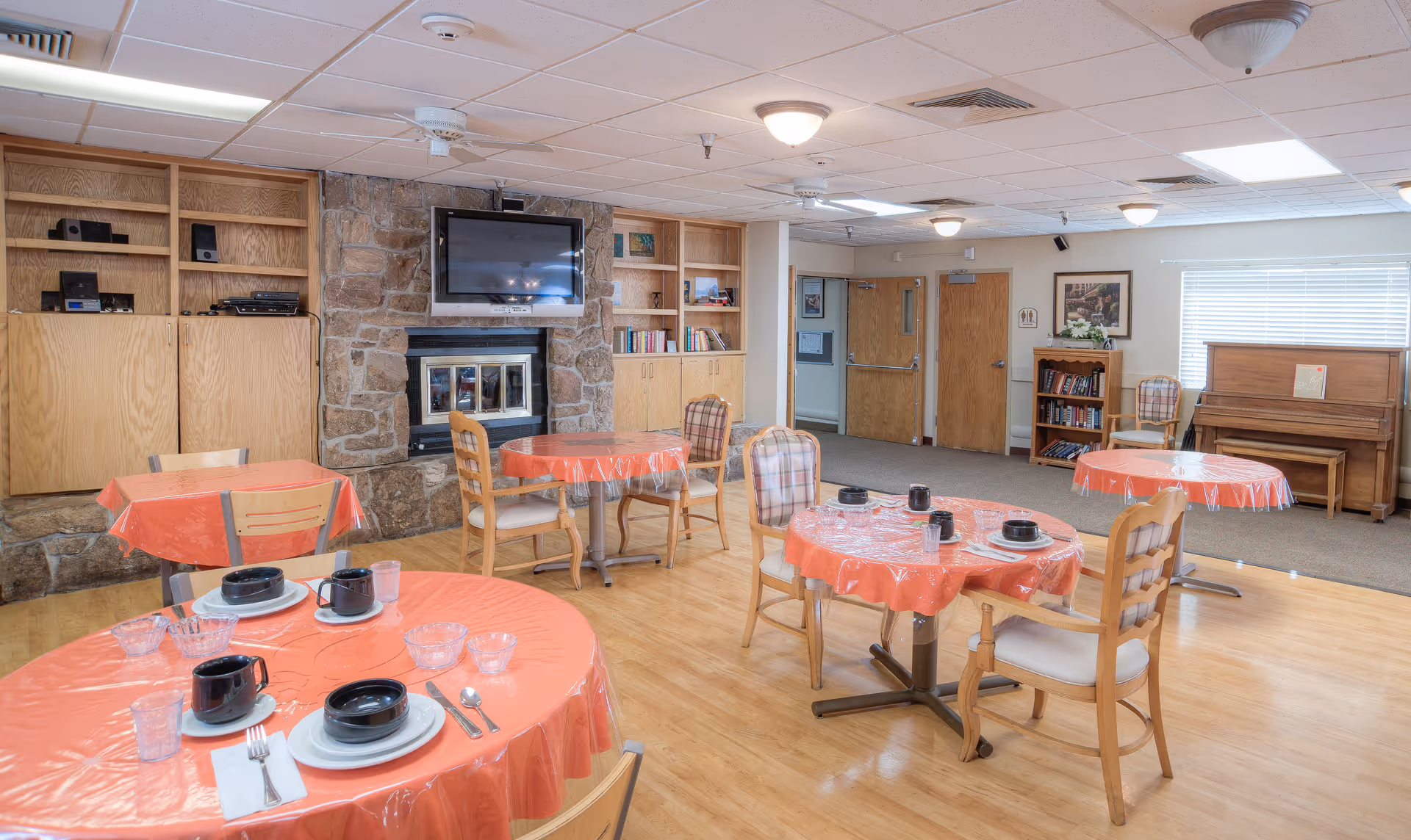 A dining area in a senior living facility with round tables covered in orange plastic tablecloths. Each table is set with black cups, bowls, plates, utensils, and clear glasses. The room features wooden chairs, a stone fireplace with a mounted TV above it, built-in wooden shelves, a piano, and a bookshelf. The floor is light wood, and there are ceiling lights and a window with blinds.
