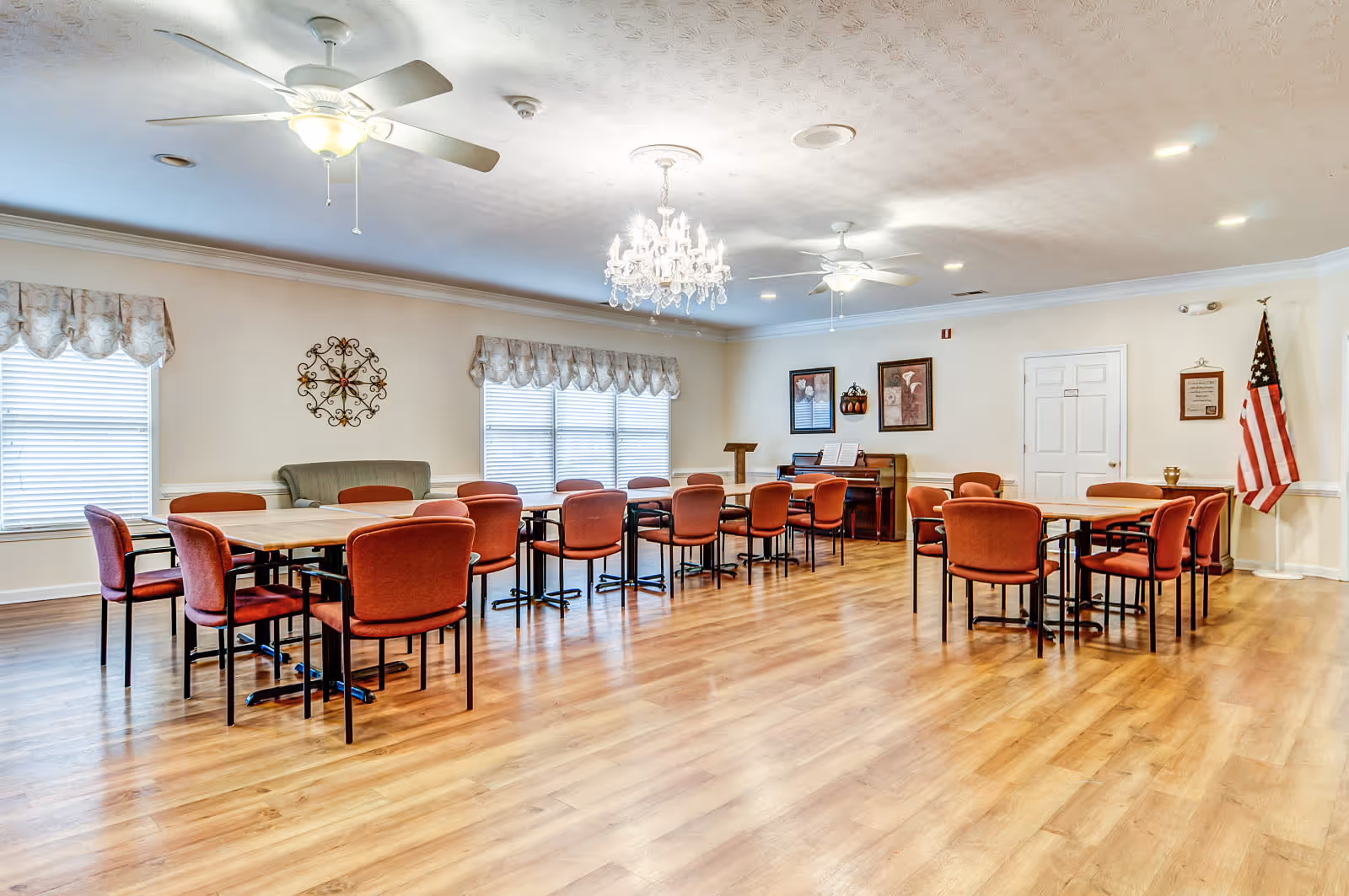 A spacious room with wooden flooring featuring several tables and red cushioned chairs arranged in groups. The room has two ceiling fans with lights, a chandelier, and large windows with valance curtains. There is a piano against the far wall with framed artwork above it, an American flag in the corner, and a green upholstered bench along one wall.