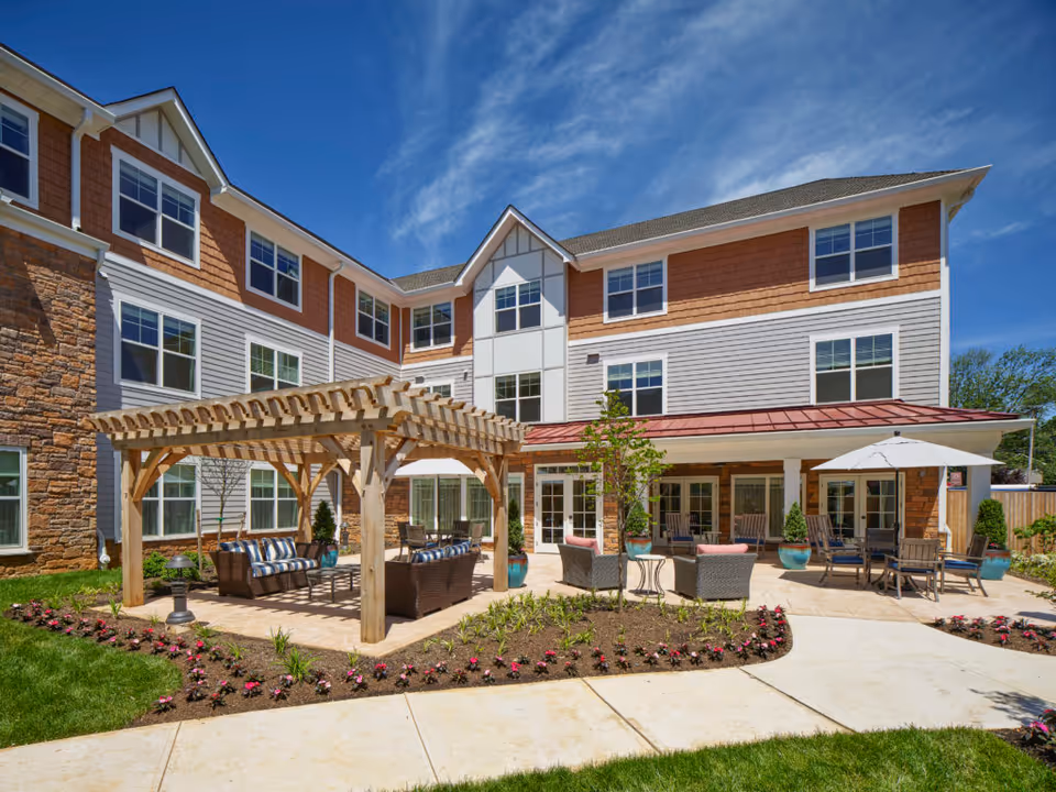 Outdoor patio area of a senior living facility with a wooden pergola covering cushioned wicker seating, additional seating with umbrellas, potted plants, and a well-maintained garden bed in front of a three-story building under a blue sky.