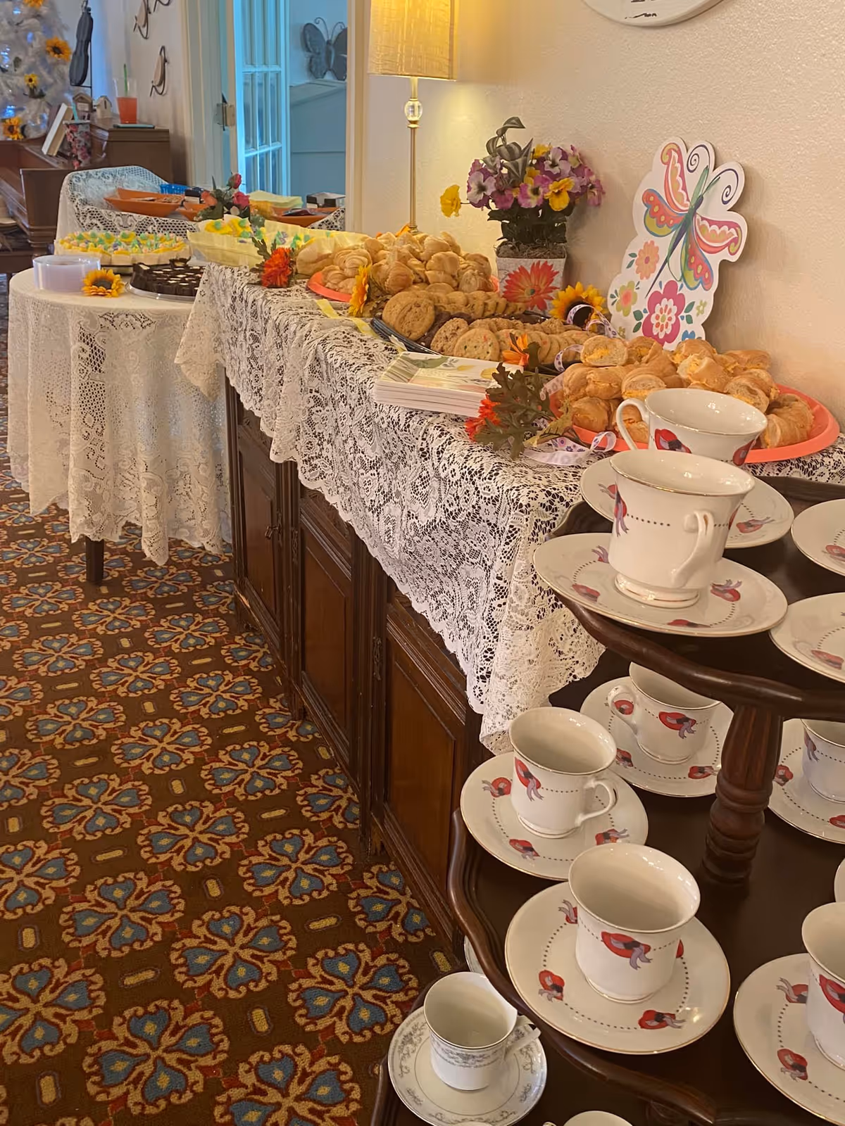 A buffet table covered with a white lace tablecloth displaying an assortment of cookies, croissants, and other pastries. Next to the table is a wooden stand holding several white teacups and saucers with red bird designs. The room has a patterned carpet and a lamp on the buffet table, along with a colorful butterfly decoration and a vase of flowers.