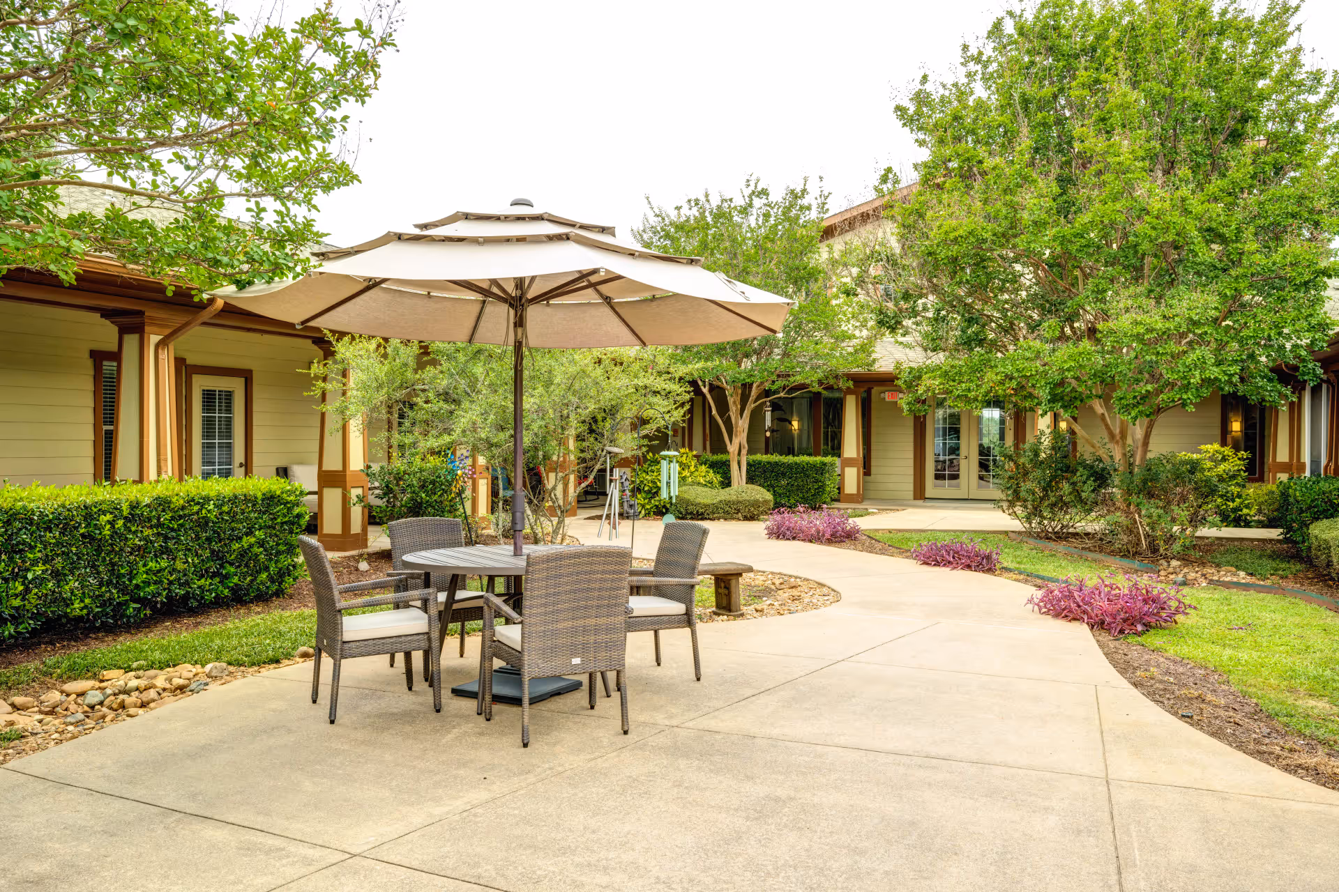Outdoor patio area at Magnolia Court Assisted Living and Memory Care featuring a round table with four wicker chairs and a large beige umbrella. The patio is surrounded by green bushes, trees, and landscaped plants with a concrete walkway leading to building entrances.