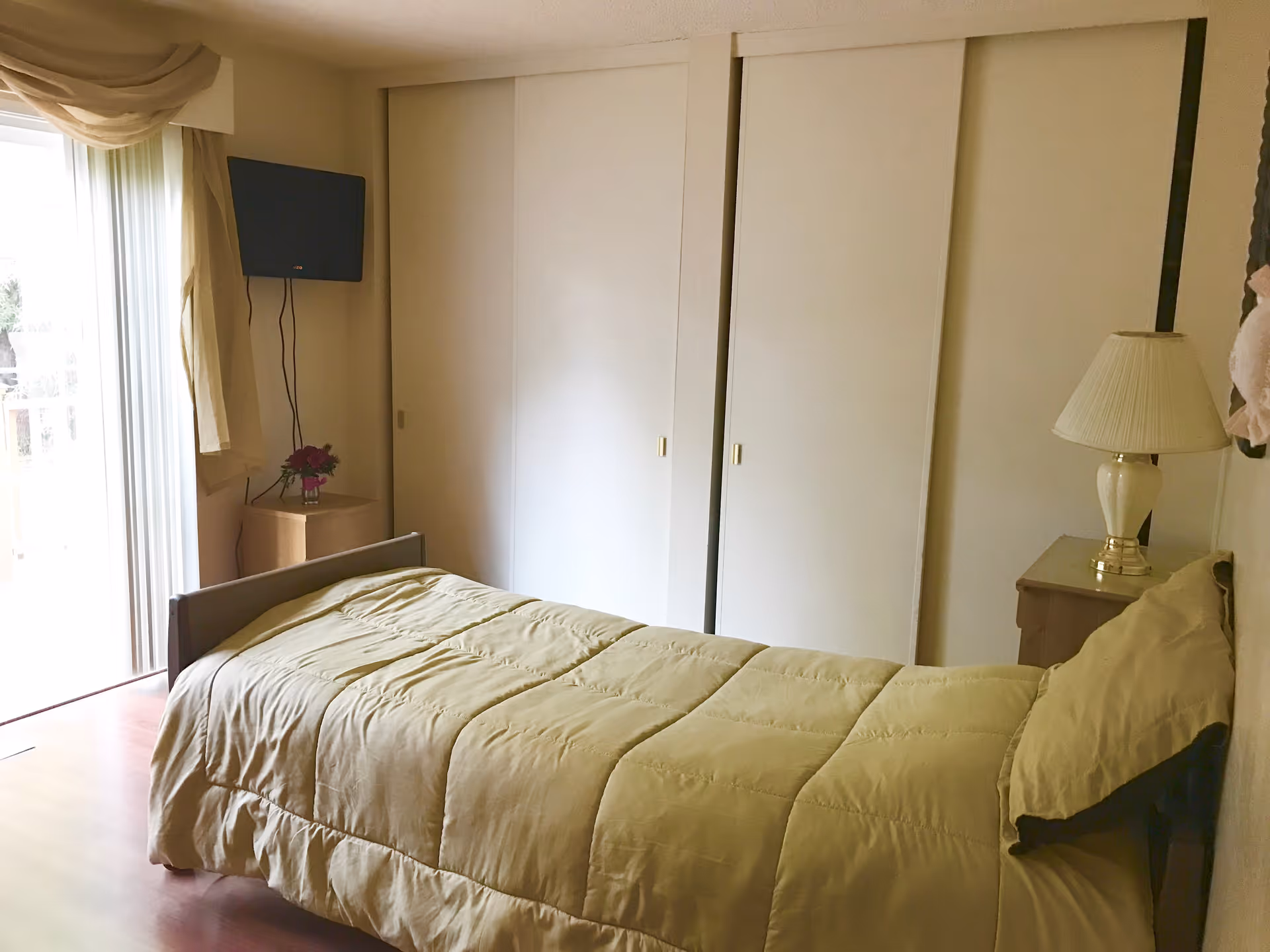 Small bedroom with a single bed, nightstand and lamp, sliding closet doors, wall-mounted TV, and a sliding glass door with curtains.