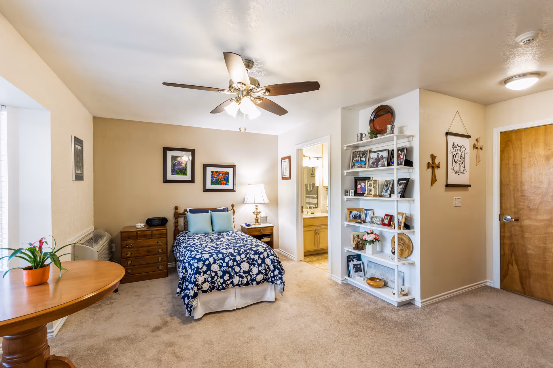 Bright furnished bedroom with a single bed covered in a blue floral comforter, a ceiling fan, shelving with framed photos, and an open doorway to a bathroom.