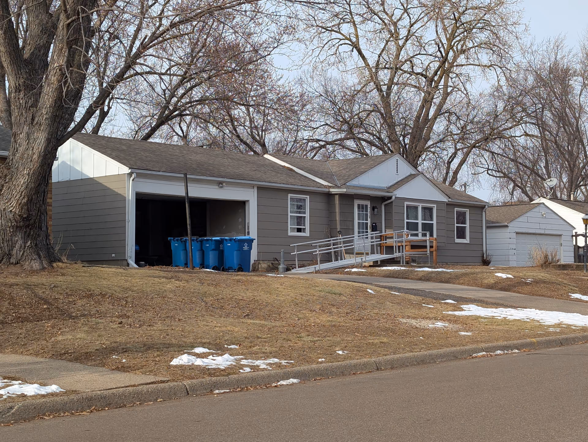 Single-story house with gray siding and a white garage door, featuring a wheelchair ramp leading to the front door. Several blue recycling bins are visible in the open garage. Leafless trees surround the house, and patches of snow are scattered on the grass and sidewalk.