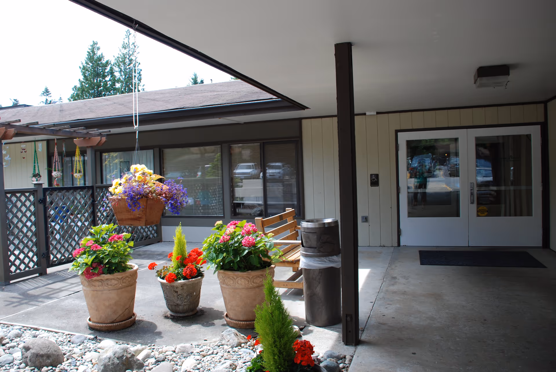 Covered outdoor entrance area of Warm Beach senior community with potted and hanging flowers, a wooden bench, a trash can, and double glass doors leading inside.