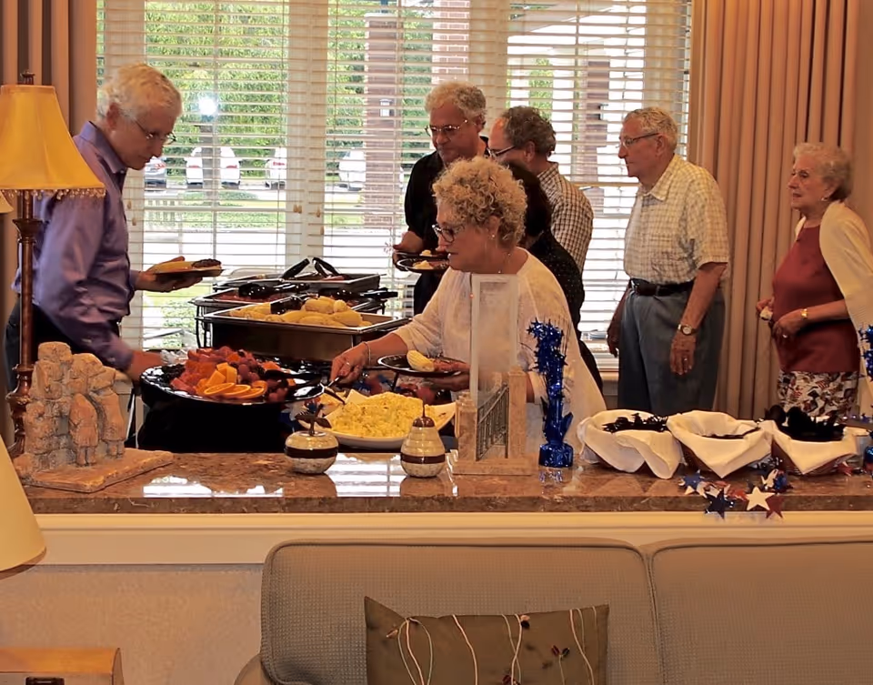 A group of elderly people serving themselves food from a buffet table in a well-lit room with large windows and beige curtains. The buffet includes fruit, scrambled eggs, and bread rolls. A lamp and decorative items are visible on the counter in the foreground.