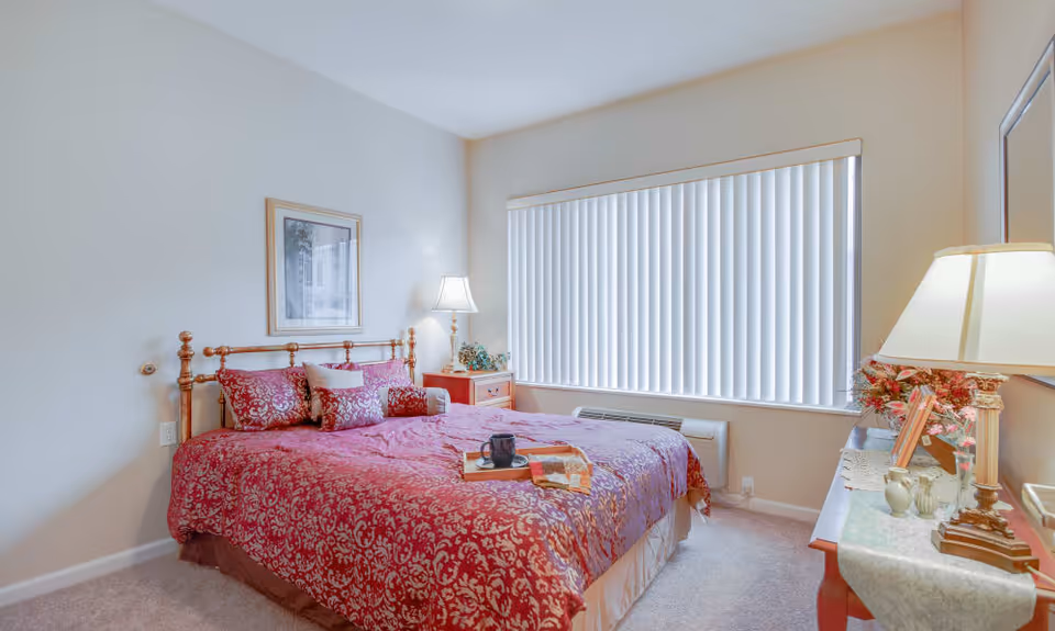 A bedroom with a large bed covered in a red patterned bedspread and multiple pillows. There is a wooden nightstand with a lamp and decorative plant on the left side of the bed. A window with vertical blinds is on the right wall. Across from the bed is a dresser with a lamp, framed photos, and floral decorations. The walls are light-colored and the floor is carpeted.