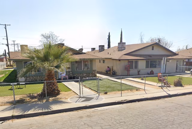 Single-story ranch-style residential building with a chain-link fence, front lawn, a palm tree, and a few people sitting near the entrance.