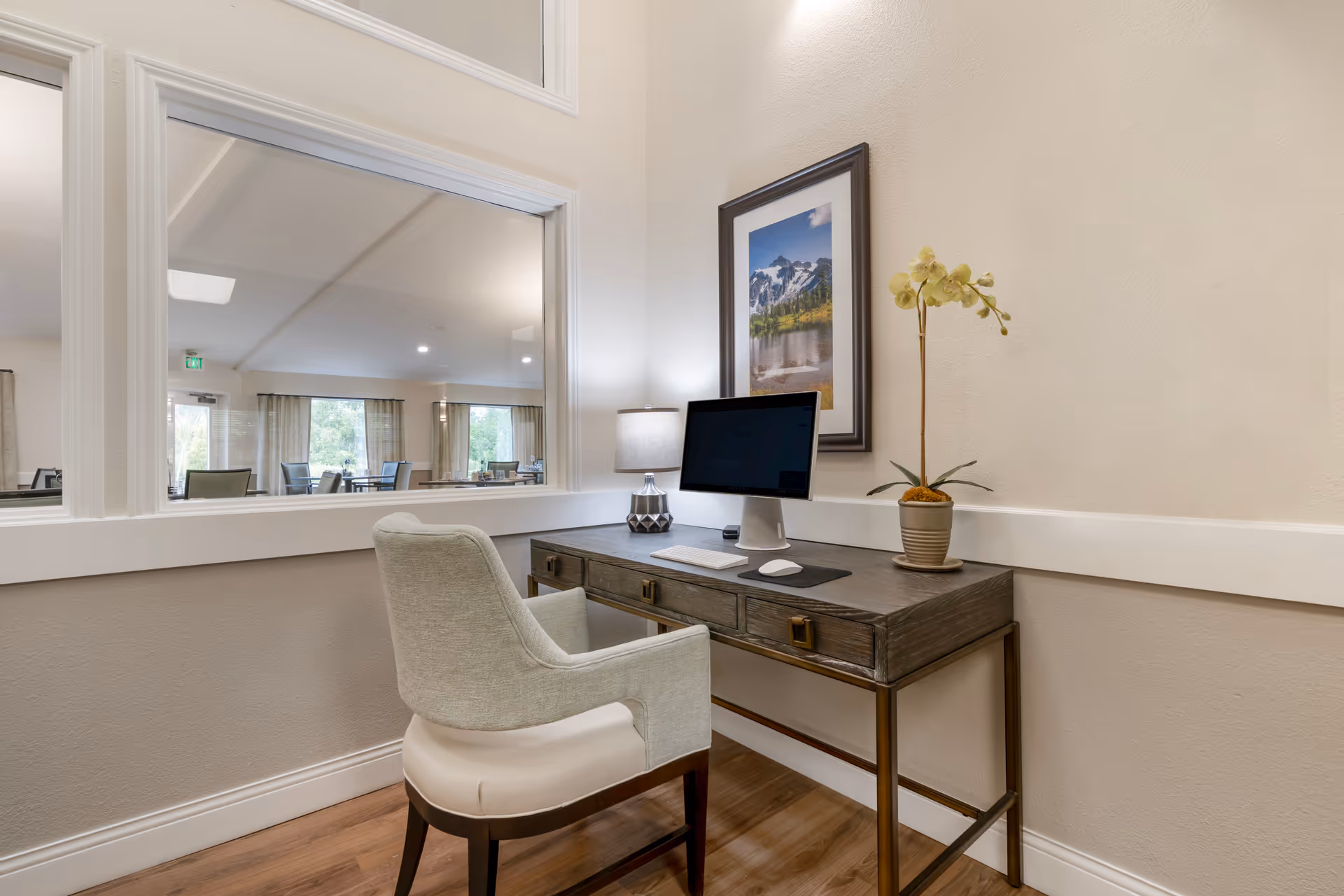 A small office nook with a wooden desk, a computer monitor, keyboard, mouse, a table lamp, and a potted orchid plant. A framed landscape photograph hangs on the wall above the desk. A large interior window looks into a dining area with tables and chairs, and large windows with curtains letting in natural light.