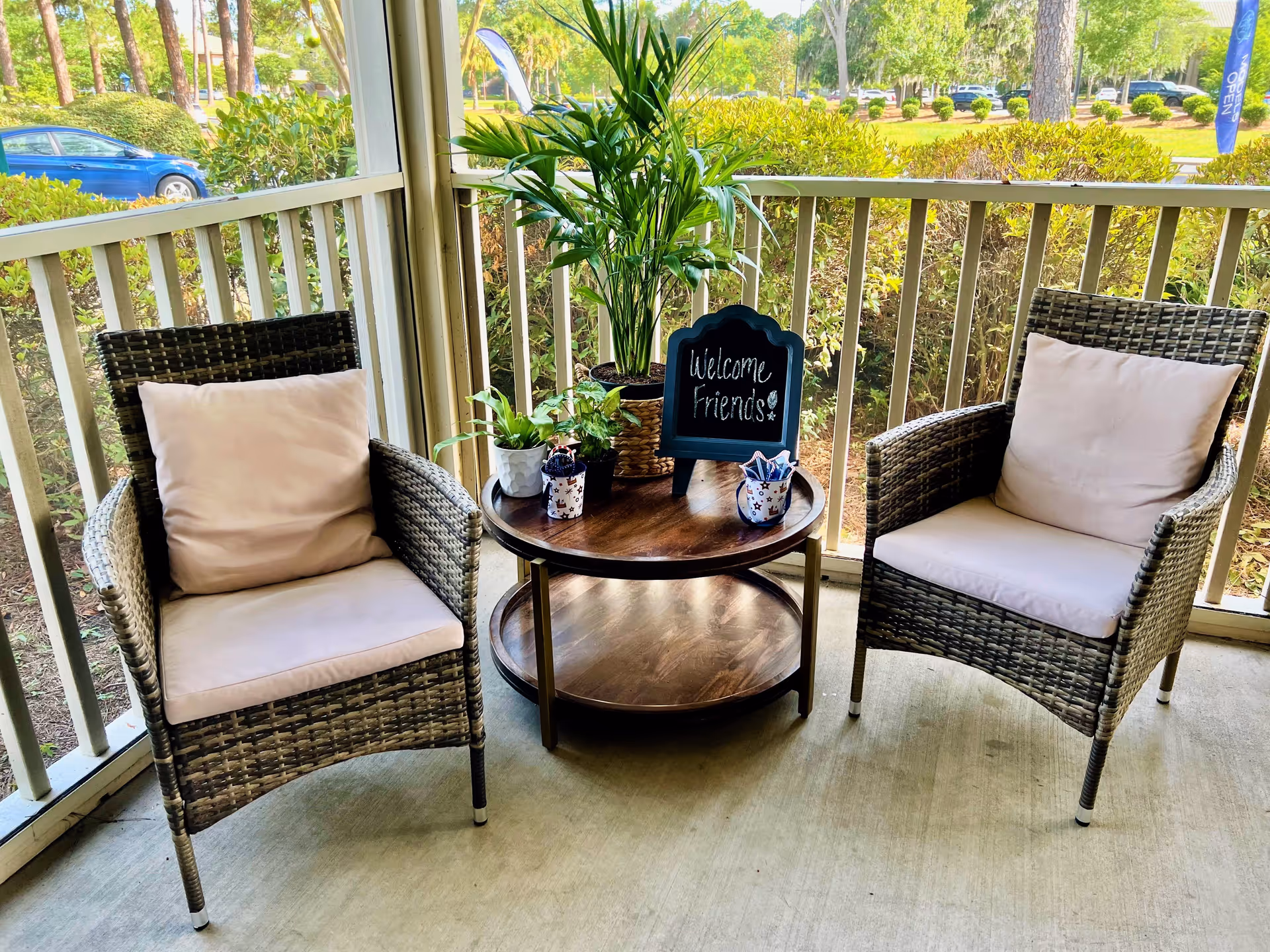 Two wicker chairs with light cushions on a covered porch or patio area. Between the chairs is a round wooden table with potted plants and a small chalkboard sign that reads 'Welcome Friends'. The porch is enclosed with white railing, and greenery and a parked blue car are visible outside.