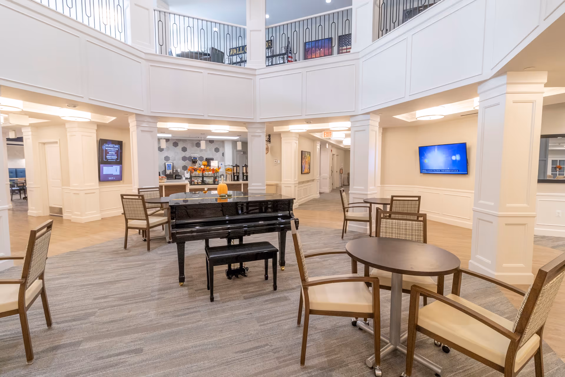 A spacious and well-lit common area in a senior living facility featuring a black grand piano in the center, several round tables with chairs, a coffee and beverage station in the background, and a TV mounted on the wall. The area has white paneled walls, carpeted flooring, and an upper balcony with railings.