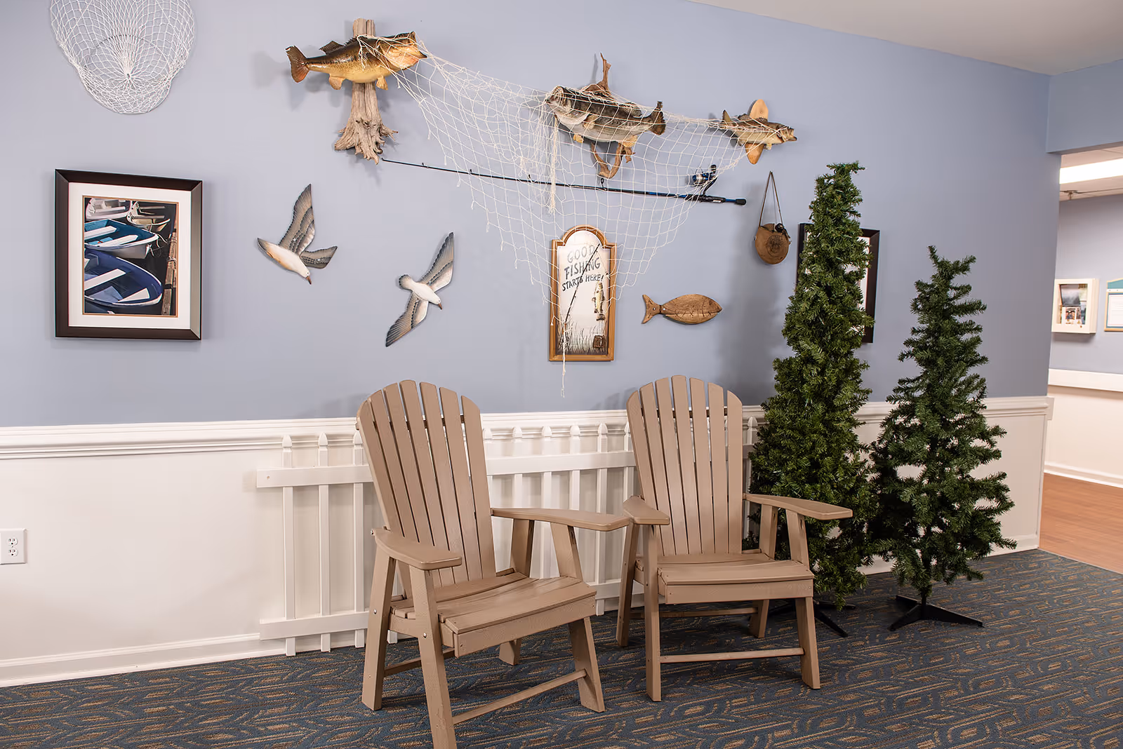 Indoor seating area with two wooden Adirondack chairs on a patterned carpet. The wall behind is decorated with fishing-themed items including mounted fish, a fishing net, wooden birds, a framed picture of boats, and a sign that reads 'Good Fishing Starts Here!'. Two artificial pine trees are placed to the right of the chairs.