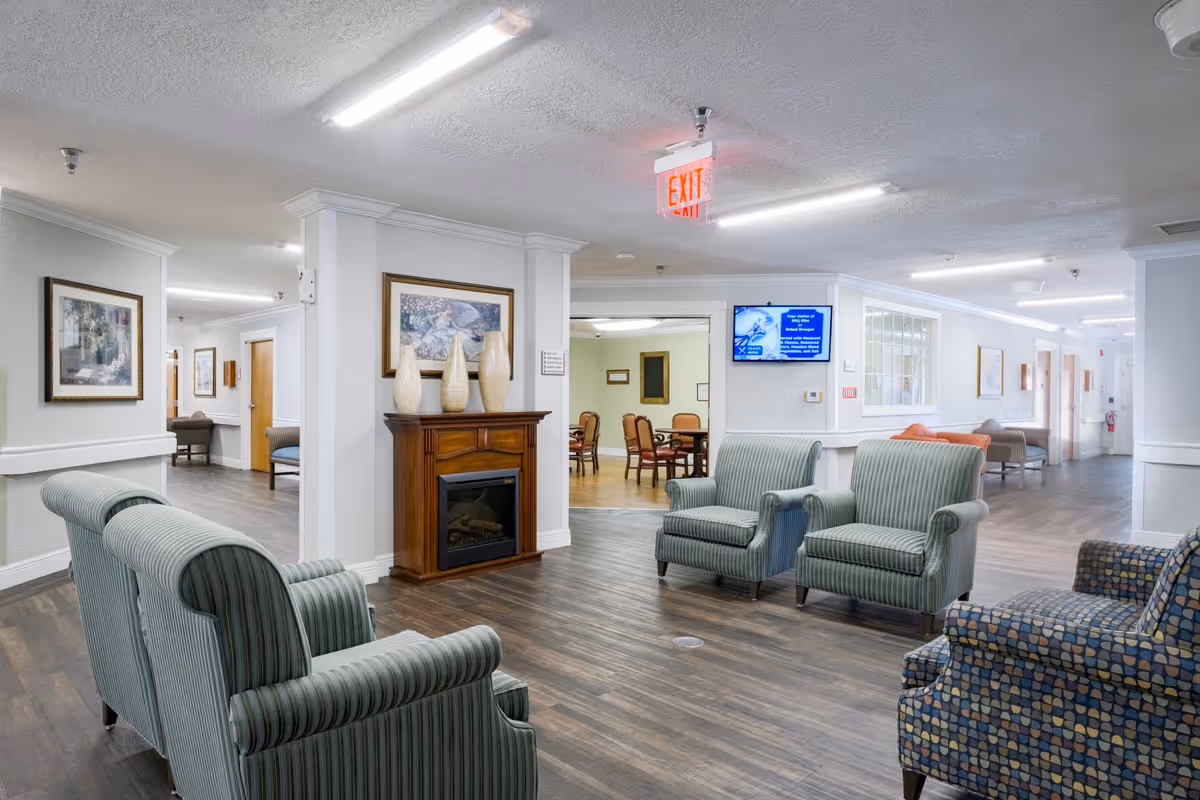 A bright and spacious senior living facility common area with several upholstered armchairs arranged around a wooden electric fireplace. The room has wood flooring, white walls, framed artwork, and a digital screen mounted on the wall. In the background, there is a dining area with tables and chairs.